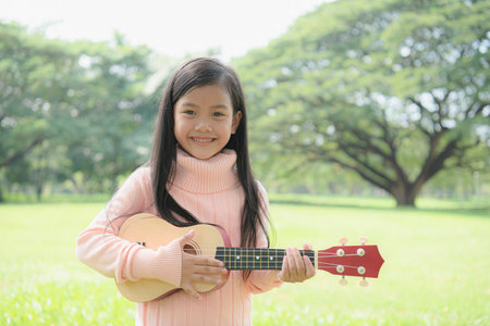 Cheerful girl playing ukulele outdoors in a green park, showcasing joy and creativity.の写真素材
