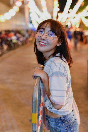 A young woman in casual attire smiles while holding onto a railing, surrounded by vibrant lights at a festive event.の写真素材