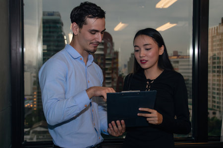 Two professionals collaborating on a digital tablet, discussing ideas in a modern office setting with city skyline view.の写真素材