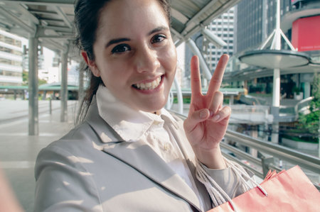 A cheerful woman in a suit enjoys urban life, holding shopping bags and flashing a peace sign outdoors.の写真素材
