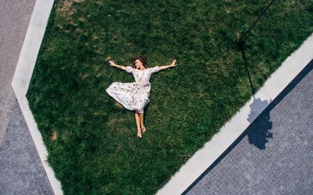 Surrealism. Happy red-haired girl lies barefoot on a green lawn in a dress. Aerial viewの写真素材