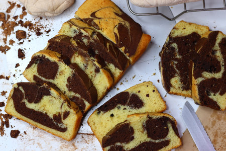 Slices of moist marble cake with chocolate and vanilla swirls, placed on a white surface. Scattered cocoa powder and chocolate shavings enhance its appeal making this image perfect for rustic dessertsの写真素材