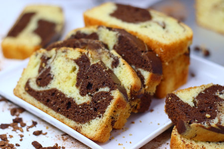 Slices of moist marble cake with chocolate and vanilla swirls, placed on a white surface. Scattered cocoa powder and chocolate shavings enhance its appeal making this image perfect for rustic dessertsの写真素材