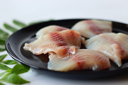 Fresh raw tilapia fillets with red veins, artfully arranged on a round black ceramic plate,...