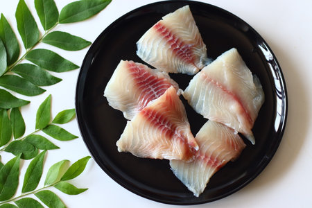 Fresh raw tilapia fillets with red veins, artfully arranged on a round black ceramic plate, surrounded with fresh curry leaves. Perfect for Asian cooking, seafood recipes, and healthy meal prepの写真素材