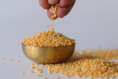 Yellow split moong dal falling into a brass bowl, with scattered lentils on white surface. A hand adds more dal, highlighting texture, color, and traditional Indian cooking ingredientsの写真素材