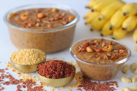 Navara rice kheer made with red rice, moong dal, jaggery, and coconut milk, garnished with fried cashews and raisins. Ingredients and bananas surround the bowls on a white surface, adding contextの写真素材
