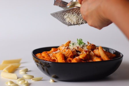Penne pasta in red sauce topped with grilled chicken, grated cheese, and parsley. A hand grates cheese over the dish. Garlic and raw pasta scattered on white surface add rustic detailの写真素材