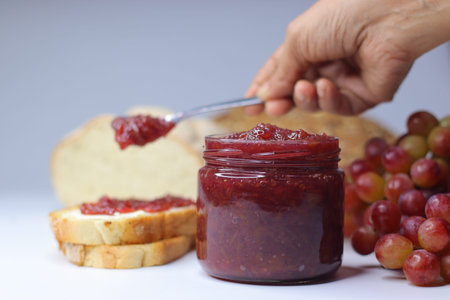 Homemade grape jam being spread on bread with a spoon, served with fresh red grapes and jam jar on...