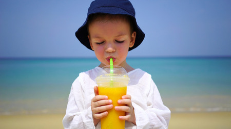 boy of two years drinking mango fresh on the beach on the background of the ocean.の写真素材