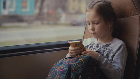 Young, beautiful girl passenger with school bag in the moving school bus using social network on her smartphone and looking out the window.の写真素材
