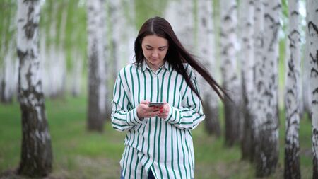 beautiful girl with a smartphone in her hands walks along a birch grove in springの写真素材