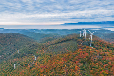 Turbine windmills for energy production. Renewable energy sources. Windmills situated on a hill overgrown with trees. Overlooking the surrounding mountains during autumnの写真素材