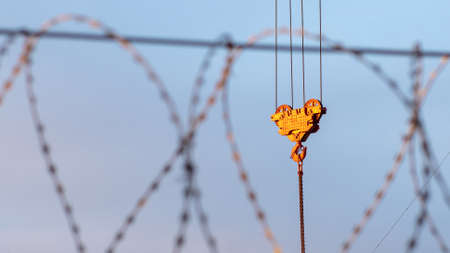 The crane's load hook is illuminated by orange rays of the sun against the sky. In the foreground is a blurred barbed wire.の写真素材