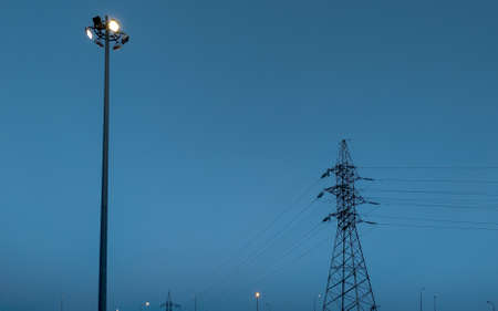 High voltage power line with a metal support and a lamppost with a luminous lamp against a blue evening sky with space for text.の写真素材