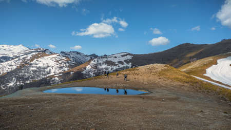 A group of tourists pass by a small reservoir in the mountains. Hiking. Mountains covered with snow. Mountain landscape.の写真素材