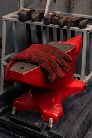 The red protective glove of the blacksmith lies on the anvil in the forge. Various hammers in the background.の写真素材