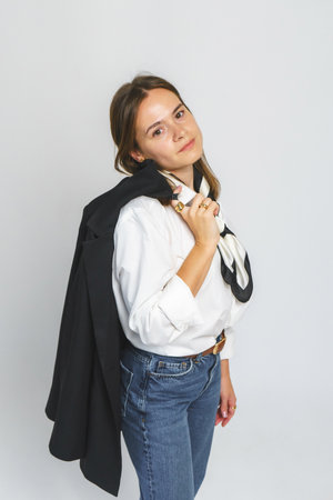 A modern businesswoman in a white shirt, blue jeans, a jacket, and a scarf on a gray background.