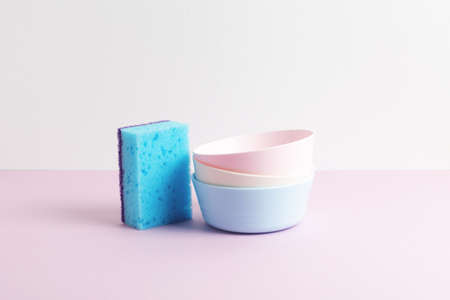 Blue sponge and colorful clean plates on pink and white background. Pastel colors. Front view. Dishwashing concept.の写真素材