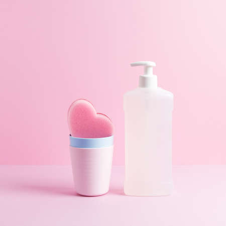 Square composition with dishwashing supplies. White bottle of dishwash liquid and pink heart shaped cleaning sponge in clean plastic cups on pastel pink background. Front view, copy space.の写真素材