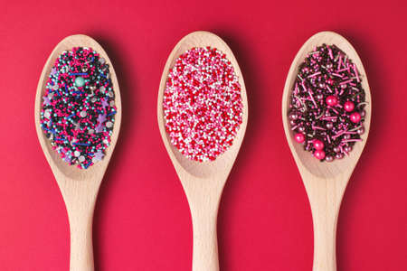 Colorful cake sprinkles in wooden spoons on red paper background. Baking decorations, top view.の写真素材