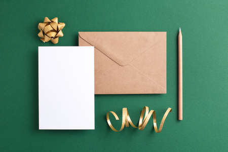 Empty card, kraft envelope, wooden pencil and golden decorations on green background. Holiday and invitation mockup.の写真素材
