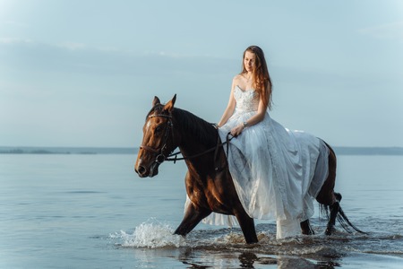 Beautiful girl in a white long dress riding a horse. Bride in the lake on horseback.の写真素材