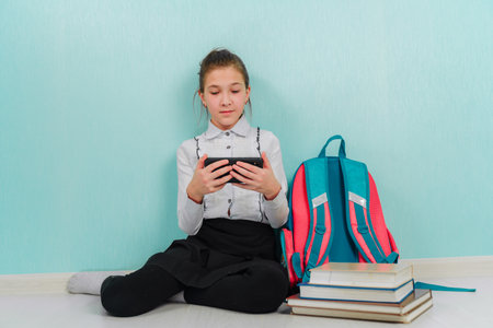 A young girl with a phone in her hands sits on the floor next to books isolated on a blue background. Distance learning at school.の写真素材
