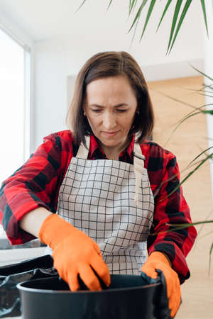 A woman in an apron pours soil into a pot for transplanting a flower. Indoor plant care.