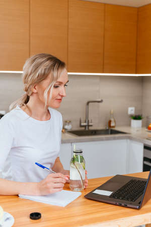 Portrait of a beautiful woman working remotely on a laptop, blonde in a white t-shirt at the table in the kitchen with a laptopの写真素材