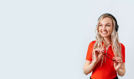 Portrait of a positive female promoter wearing red glasses and headphones. Happy girl in a red T-shirt, isolated on a white background, holds glasses in her handsの写真素材