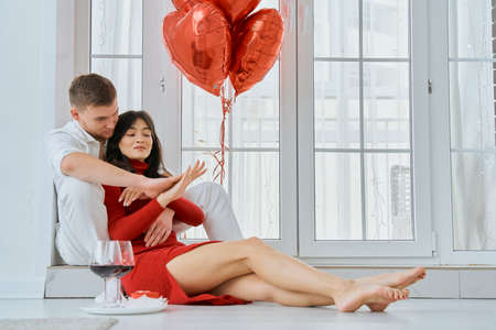 A loving couple sits near the window in an embrace. A girl in a red dress with her beloved boyfriend. Engagement. Valentines Day.の写真素材