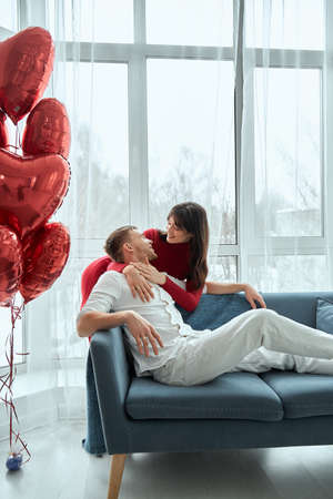 The girl gently hugs the guy sitting on the couch. Valentines Day concept. Happy couple in love. The couple celebrates Valentines Day at home. Romantic home for Valentineの写真素材