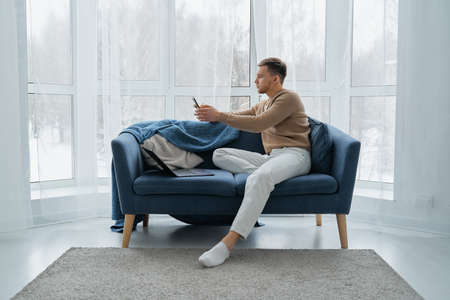 A man with a mobile phone and a laptop is sitting on a blue sofa against the background of a window. A young man works at home on the couch with his laptopの写真素材