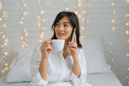 a girl with a cup of tea sits on the bed and talks on the phone.
