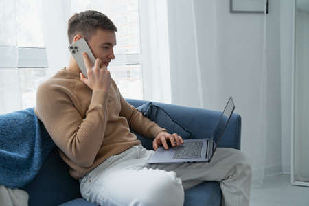 A young man works at home on the couch with his laptop. A man is talking on a mobile phone while sitting on a blue sofa with a laptop.の写真素材
