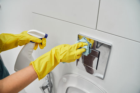 A womans hand in yellow rubber gloves wipes the toilet flush button with a rag. Sanitary treatment in the bathroom.の写真素材