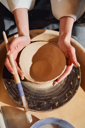 Close-up of female hands with a brush in hand with a clay bowl. The potter works in his workshop on the potters wheel and makes clay productsの写真素材
