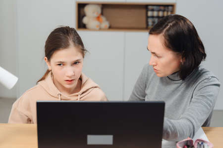 A happy family. Girl and mother are doing homework while sitting at a laptop in the room. Mom teaches daughter sitting at the table at home.の写真素材