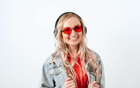 Blonde in headphones on a white background. She is wearing a denim jacket and red glasses.の写真素材