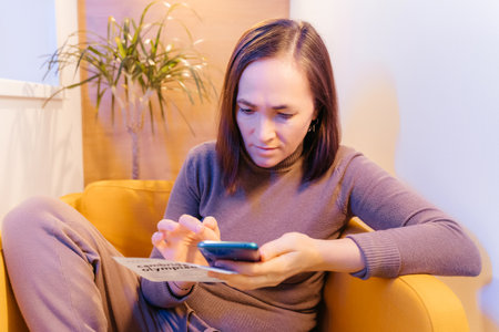 A woman is resting in a cozy soft chair with a smartphone. Pretty smiling woman is looking at the screen of the device, and is typing. Social media funの写真素材