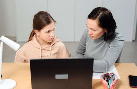 A happy family. Girl and mother are doing homework while sitting at a laptop in the room. Mom teaches daughter sitting at the table at home.の写真素材