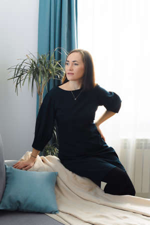 Happy relaxed young woman sitting on sofa armrest in living room at home. Portrait of a girl in a blue dress against the background of a green plant and curtains.の写真素材