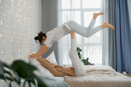 Lying on a comfortable bed, a man raises his girlfriend with his feet, as if floating in the air. A young couple in love play, spend their free time, having fun at home, in the bedroom.の写真素材