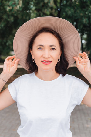 middle-aged woman in a summer hat looks at the camera, posing against the backdrop of trees.の写真素材