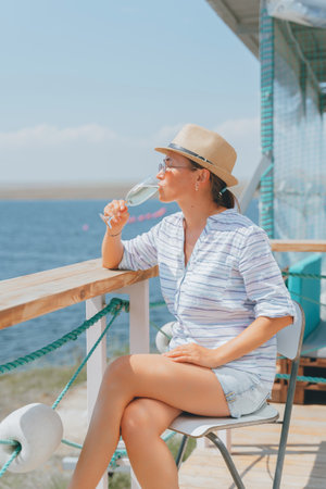 A woman in a straw hat and sunglasses sits in a restaurant with a glass of champagne overlooking the sea.の写真素材