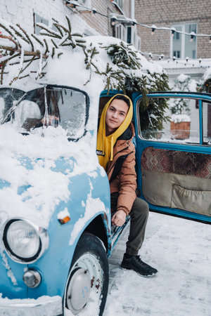 A young man sits on the seat of a vintage car with the door open, a freshly cut down Christmas tree on the roof of a vintage car. Christmas.の写真素材