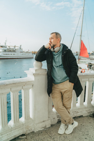 Pensive middle-aged man in headphones and a black jacket in a seaport against the backdrop of yachts.の写真素材