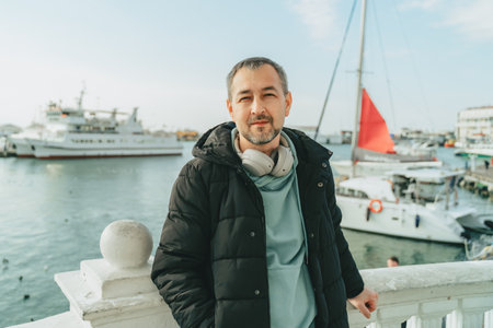 A middle-aged man in a black jacket and full-size headphones in front of yachts.の写真素材