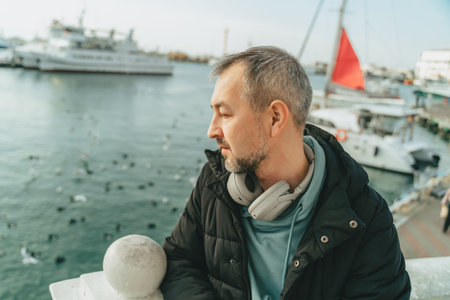 A middle-aged man in a black jacket and full-size headphones in front of yachts.の写真素材